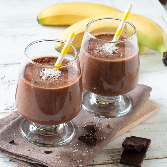 Two glasses of Mexican Chocolate Protein Shake with straws on a table with bananas and chocolate. 