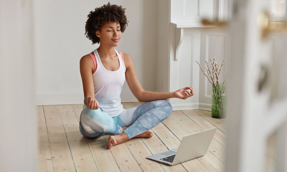 Black woman doing yoga in Easy Pose (Sukhasana) on the floor with her laptop.